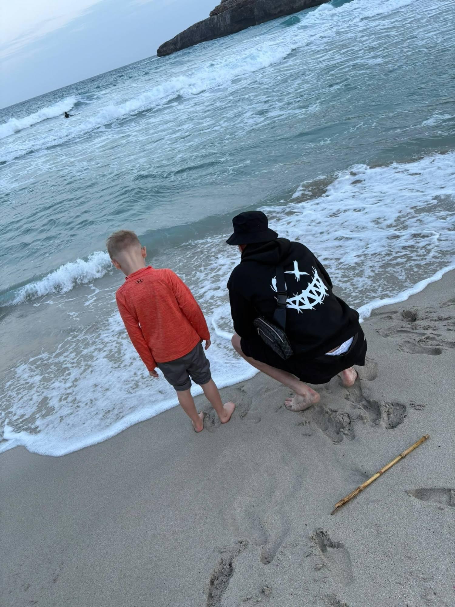An adult crouching beside a young boy at the water's edge on a beach, both seen from behind.